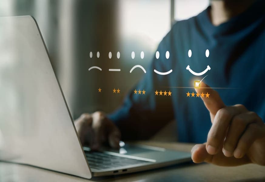 A person sitting at a desk with a laptop in front. Floating on the image is a scale of smiley faces ranging from a frowny face to a happy face with corresponding star ratings underneath ranging from 1 to 5