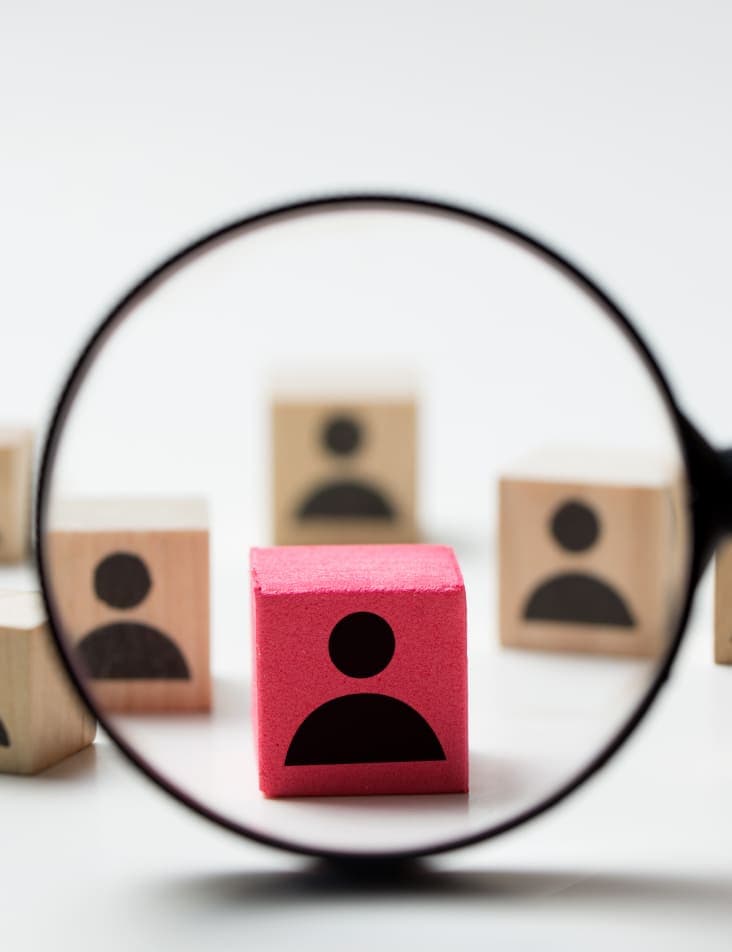 A cluster of wooden blocks with very basic human silhouettes printed on them and a magnifying glass focusing on just one that is painted red